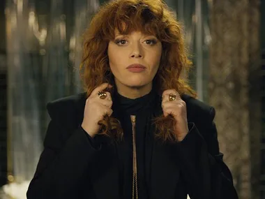 A woman with curly red hair wears a black outfit, standing confidently with her hands near her neck.