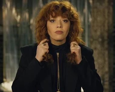 A woman with curly red hair wears a black outfit, standing confidently with her hands near her neck.