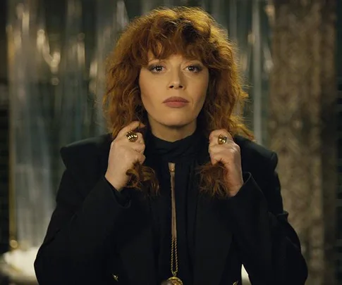 A woman with curly red hair wears a black outfit, standing confidently with her hands near her neck.
