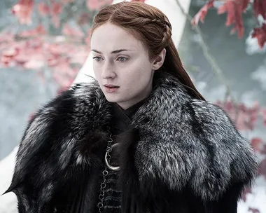 Sansa Stark in a fur coat stands amid autumn leaves in "Game of Thrones" scene.