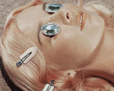 Woman with pink hair and gem-like glasses lies on the ground, reflecting light and wearing hair clips.