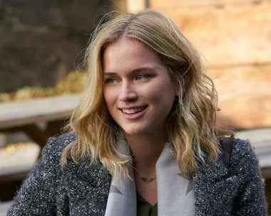 A woman with blonde hair smiles, wearing a textured coat and scarf, sitting outdoors on a wooden bench.