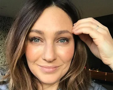 A woman with shoulder-length brown hair and blue eyes smiles gently while touching her eyebrow.