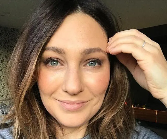 A woman with shoulder-length brown hair and blue eyes smiles gently while touching her eyebrow.