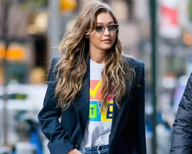 Woman in sunglasses wearing a navy blazer and graphic T-shirt, walking in an urban setting with wavy hair loose.