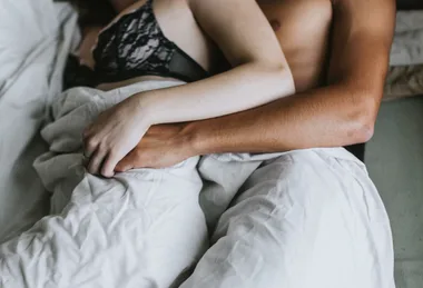 Couple lying in bed, partially covered with a white sheet, holding hands.