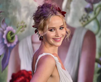 Actress in a glamorous white dress with floral backdrop, smiling at a public event.