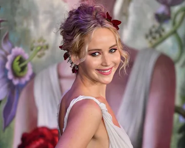 Actress in a glamorous white dress with floral backdrop, smiling at a public event.