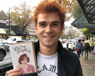 Person with orange hair smiling, holding a card that reads "you're pretty," standing on a street with parked cars and trees.