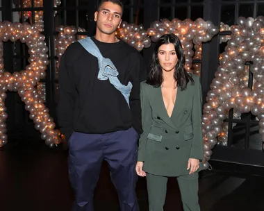 A man in a black sweater and pants stands next to a woman in a green suit with balloon decorations in the background.