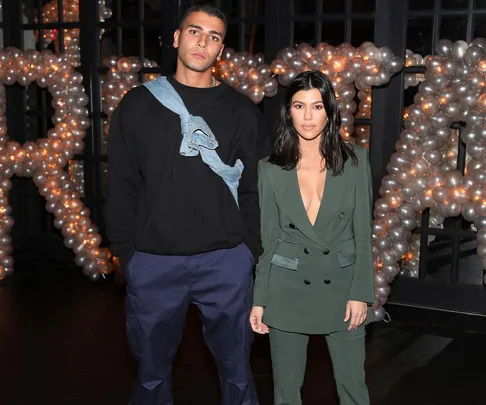 A man in a black sweater and pants stands next to a woman in a green suit with balloon decorations in the background.