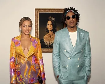 Beyoncé and Jay-Z pose in colorful outfits beside a framed portrait of Meghan Markle wearing a crown and jewelry.