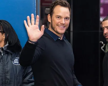 Man in dark sweater waving and smiling at the camera, standing near a security staff member.