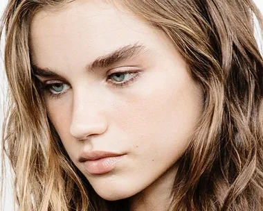 Young woman with thick eyebrows and wavy brown hair looking down thoughtfully.