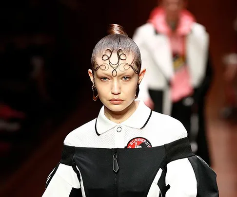 Model on runway in a Burberry outfit with intricate hairstyle and badge, walking with serious expression; blurred background.