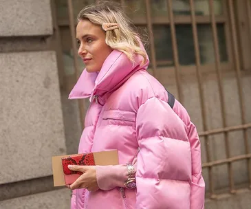 Woman in pink puffer jacket with a hair clip, holding a red clutch, walking outdoors.