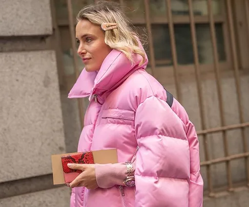 Woman in pink puffer jacket with a hair clip, holding a red clutch, walking outdoors.