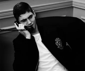 Young man in a casual jacket, sitting and talking on a corded phone, expression curious or amused, in black and white.