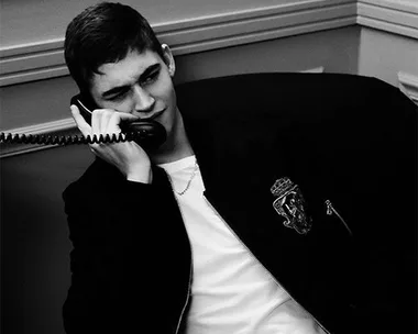 Young man in a casual jacket, sitting and talking on a corded phone, expression curious or amused, in black and white.