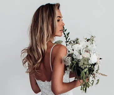 Bride in lace dress holding large bouquet of white flowers against minimal background.