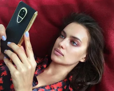 Woman lying on a red blanket, looking at a smartphone, with light purple nail polish and a red-patterned top.