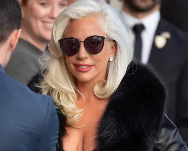 Lady wearing sunglasses and a fur coat, smiling outside with people in the background.