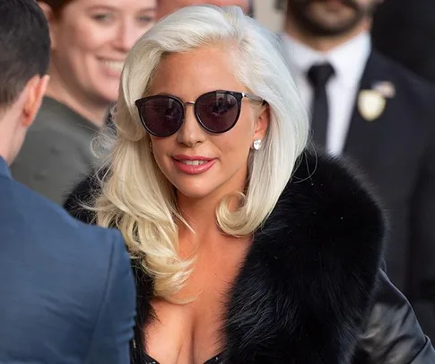 Lady wearing sunglasses and a fur coat, smiling outside with people in the background.