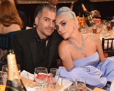 Lady Gaga and Christian Carino seated together at an event, surrounded by glasses and a bottle on the table.