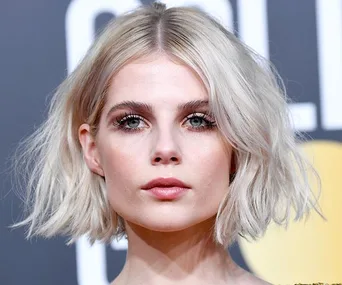 Woman with short, wavy blonde bob hairstyle, wearing makeup, in front of a blurred gray background.