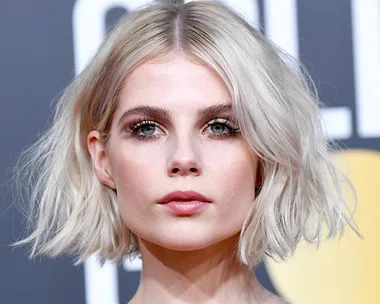 Woman with short, wavy blonde bob hairstyle, wearing makeup, in front of a blurred gray background.