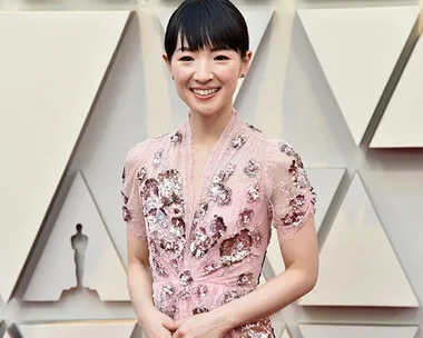 Smiling woman in a pink floral gown at the Oscars 2019 red carpet with geometric pattern backdrop.