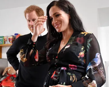 Pregnant woman in floral dress smiles and gestures, with a man standing alongside in a casual setting.