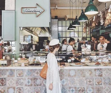 Woman in white dress and beret choosing from pastries at a bustling café counter.