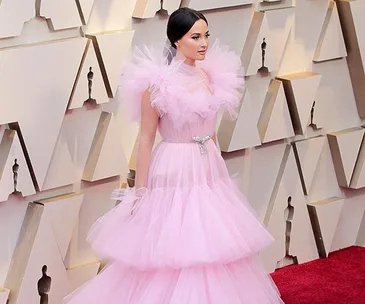 Person in a voluminous pink tulle dress on the Oscars 2019 red carpet, with geometric background.