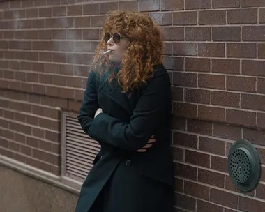 Woman with curly hair, wearing sunglasses, smokes against a brick wall.