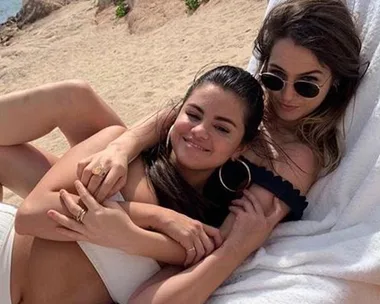 Two women smiling and relaxing on a beach lounge chair, one wearing sunglasses, both in swimwear.