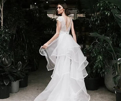 A bride in a layered white wedding gown stands amidst green plants, looking over her shoulder in an indoor setting.