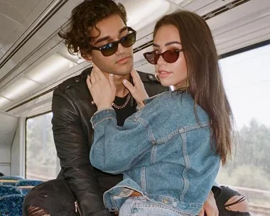 Couple wearing sunglasses pose closely on a train, both in denim jackets.