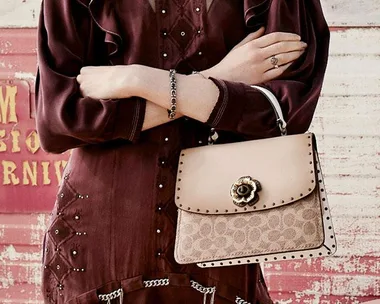 A person in a maroon dress holds a beige Coach handbag with a floral clasp against a textured backdrop.