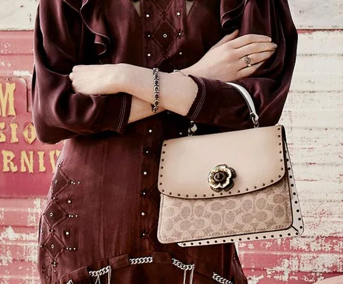 A person in a maroon dress holds a beige Coach handbag with a floral clasp against a textured backdrop.