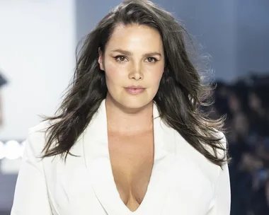 A woman with long brown hair in a white outfit walking on a runway, focused expression.