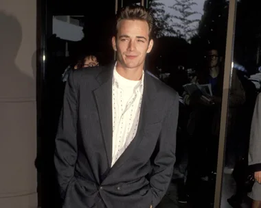 Man in a suit and dotted shirt, hands in pockets, standing by a reflective glass, evening setting.