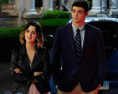 Two characters from "The Perfect Date" stand beside a car, dressed formally, with trees and a building in the background.