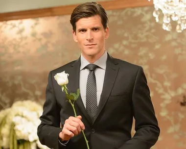 Man in a suit holding a white rose in an elegant room with a floral background.