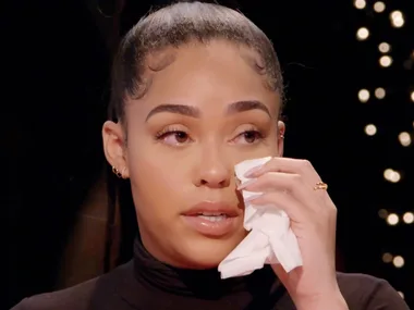 Woman with tied-back hair, holding tissue to her face, appears emotional during an interview against a softly lit background.