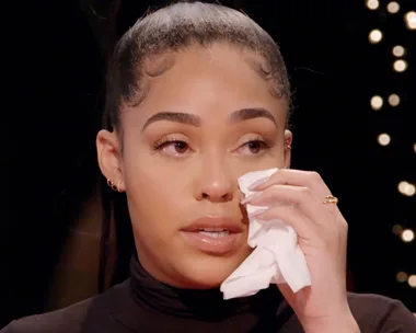 Woman with tied-back hair, holding tissue to her face, appears emotional during an interview against a softly lit background.