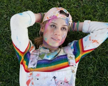 Person with a rainbow sweater and paint-splattered overalls lies on grass, looking thoughtful.