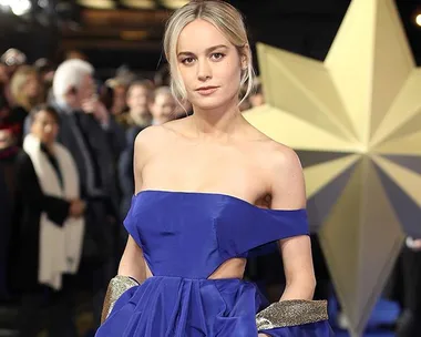 A woman in a blue dress poses confidently at a glamorous event with a large star decoration and crowd in the background.