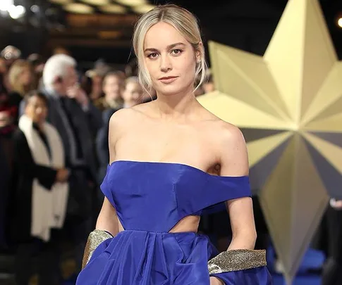 A woman in a blue dress poses confidently at a glamorous event with a large star decoration and crowd in the background.