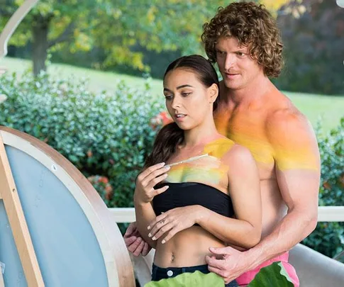 A man and woman are body painting outdoors, both topless and covered in colorful paint.